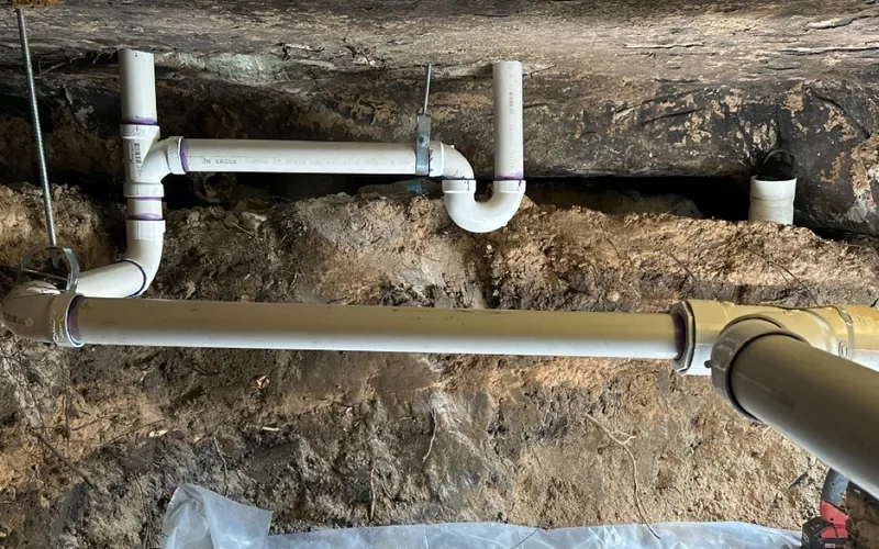 A tunnel dug beneath a house slab, the underside of the foundation visible along the top, with a new white PVC drain line and trap assembly run through it.